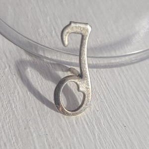 Pode incluir: Um pingente prateado em forma de nota musical. O pingente tem um topo curvo e uma laçada na parte inferior com um recorte em forma de coração. É apresentado em um colar fino e transparente sobre um fundo branco.