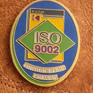 Op de afbeelding: Een vintage ovale speld met een gouden rand. De speld heeft een blauwe achtergrond met een groene ruit in het midden, met de tekst "ISO 9002". De bovenkant leest "Kodak". De onderkant leest "FINITION RADIO CHALON".
