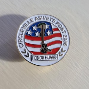 May include: A gold-coloured pin with a white background and red and blue stripes. The pin features a soldier silhouette standing in front of an American flag with the text "CIRCLEVILLE AMVETS POST 2256 HONOR GUARD".