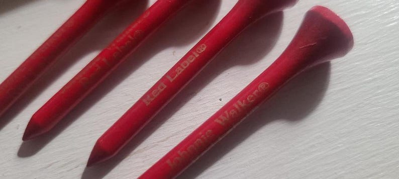 Może przedstawiać: Zbliżenie czterech czerwonych kołk&oacute;w golfowych. Kołki mają zaokrągloną g&oacute;rę i spiczaste dno. Na kołkach wydrukowano tekst "Red Label" i "Johnnie Walker".