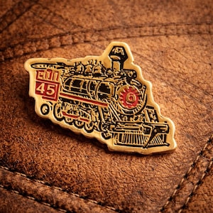 May include: A gold-colored train pin with black and red accents. The pin features a detailed illustration of a vintage steam engine, with the number "45" in a red box. The pin is set against a brown leather background.