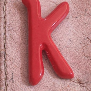 May include: A glossy, bright red letter "K" rests on a textured, pinkish-brown surface. The letter is a solid, three-dimensional shape with rounded edges, suggesting a decorative or craft item.