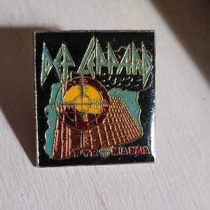 Könnte beinhalten: Quadratischer Metall-Pin mit dem Def Leppard-Logo in Teal und Gold, mit einem Ziel- und Pyramiden-Design. Das Wort "PYROMANIA" steht unten. Der Pin hat einen schwarzen Hintergrund und ein glänzendes Finish.