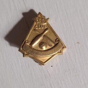 May include: A vintage gold-colored bowling pin with a bowling ball and the words "500 CLUB" on a triangular pin. The pin has a banner at the bottom and is likely a bowling achievement award.