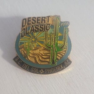 May include: A gold-colored pin with the words "DESERT CLASSIC" and "2006" in gold lettering. The pin features a desert landscape with cacti and a sun design. The bottom of the pin reads "S.C. DEL SOL - PHOENIX, AZ".