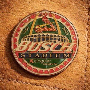 May include: A round, colorful pin featuring the Busch Stadium logo and the St. Louis Cardinals logo. The pin has a red, green, and gold color scheme, with the years 1966 and 2005 on the edges. The text "singular, raising the bar" is also visible.
