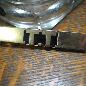 May include: A gold-tone tie bar with black enamel letters spelling "TTT".