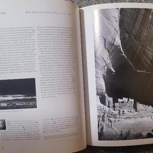 May include: An open book with two pages. The left page has text and a small black and white photo. The right page features a large black and white photograph of a cliff dwelling. The book is open on a patterned surface.