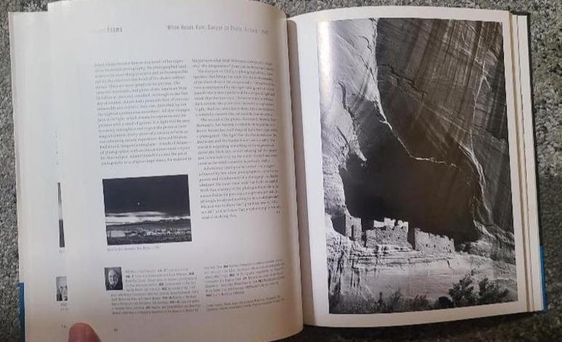 May include: An open book with two pages. The left page has text and a small black and white photo. The right page features a large black and white photograph of a cliff dwelling. The book is open on a patterned surface.