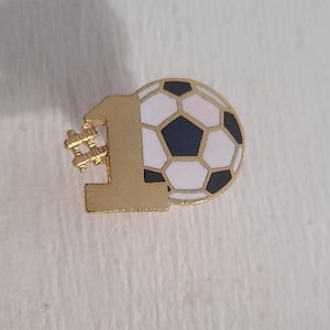 May include: A gold-colored pin featuring the number 1 with a hashtag symbol and a soccer ball design. The soccer ball has white, black, and navy blue pentagons and hexagons. The pin is likely a sports-themed accessory.