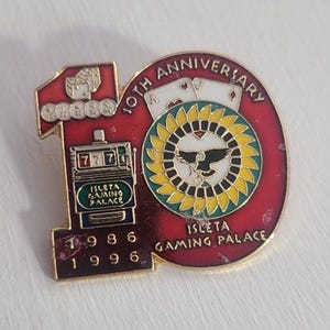 May include: A gold and red pin commemorating the 10th anniversary of the Isleta Gaming Palace. The pin features a slot machine, playing cards, and a stylized sun with a bird inside. The text "10th Anniversary 1986 1996" is printed on the pin.