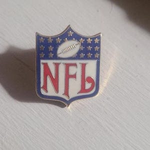 May include: A metal NFL pin with a shield shape. The pin features a blue background with white stars, a white football, and the letters "NFL" in red. The pin has a gold border.