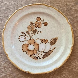Può includere: Un piatto rotondo in ceramica bianco sporco con bordo smerlato. Il piatto presenta un motivo floreale nei toni del marrone e del bianco. I fiori sono raggruppati al centro del piatto. Il bordo del piatto ha un bordo marrone.
