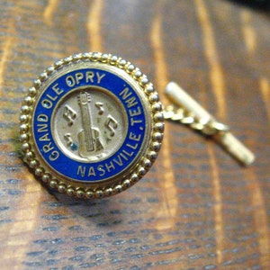 May include: Gold-coloured tie tack with a blue enamel circle featuring the text "GRAND OLE OPRY NASHVILLE, TN." and a gold-coloured guitar with musical notes.