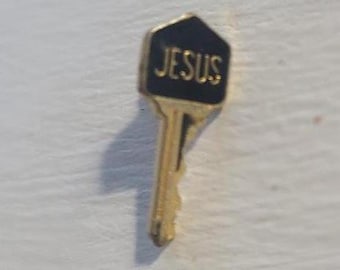 May include: A gold-colored key-shaped pin with a black, hexagonal top. The word "JESUS" is written in gold lettering on the black top. The pin is set against a white background.