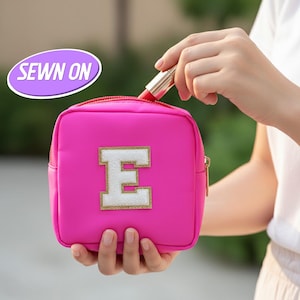 Puede incluir: Un neceser rosa brillante con cremallera dorada y una gran letra "E" blanca con ribete dorado brillante. La bolsa tiene las palabras "SEWN ON" en un óvalo púrpura. Una persona sostiene la bolsa y un lápiz labial.