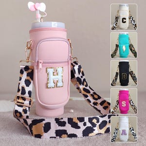 Könnte beinhalten: Ein rosa Wasserflaschenträger mit Leopardenmuster-Gurt und goldfarbener Hardware. Der Träger hat eine Vordertasche mit dem weißen und goldenen Buchstaben "H". Zusätzliche Wasserflaschen mit verschiedenen Farben und Buchstaben sind ebenfalls abgebildet.