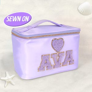 Peut inclure: Trousse de maquillage lilas avec une poignée, une fermeture éclair dorée et le nom "AVA" en lettres violettes avec des bordures dorées. Un cœur violet avec des paillettes dorées est au-dessus du nom. Le texte "SEWN ON" est dans un ovale violet.