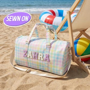 Op de afbeelding: Een pastelkleurige geruite duffeltas met de naam "ZAHRA" geborduurd op de voorkant. De tas heeft een schouderriem en handvatten aan de bovenkant. Een strandscène met een strandstoel, opblaasbare ring en bal is op de achtergrond.