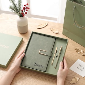 May include: A sage green notebook with a gold-toned rectangular clasp and the name Teresa printed in black, paired with a matching pen in a gift box. The set includes a sage green gift bag and a small white card with the text With Love.