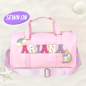 Puede incluir: Bolsa de deporte rosa con el nombre "ARIANA" en letras coloridas cosidas. La bolsa presenta un parche de arcoíris y unicornio, una correa para el hombro lavanda y una hebilla dorada. El texto "SEWN ON" está en un óvalo morado.