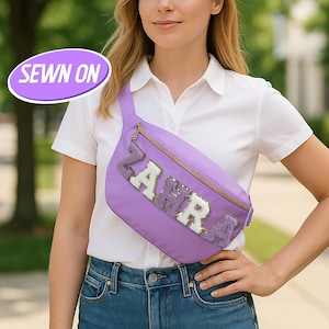 May include: A lavender fanny pack with a gold zipper and the name "ZAHRA" spelled out in sequined letters. The fanny pack is worn across the body. The words "SEWN ON" are in a purple oval in the upper left corner.