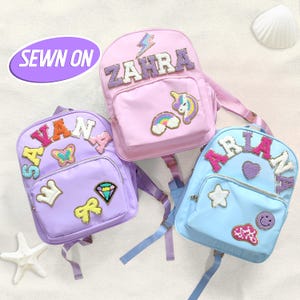 Peut inclure: Trois sacs à dos colorés personnalisés avec des noms et des écussons décoratifs. Les sacs sont de couleurs violet, rose et bleu. Chaque sac présente un nom épelé en lettres à paillettes, ainsi que divers écussons brodés tels que des licornes, des arcs-en-ciel et des étoiles.