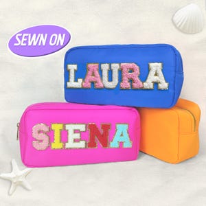 Pode incluir: Três nécessaires coloridas. Uma bolsa azul com o nome "LAURA" em letras rosa e brancas, uma bolsa rosa com "SIENA" em letras coloridas e uma bolsa laranja. As bolsas estão sobre uma superfície arenosa.