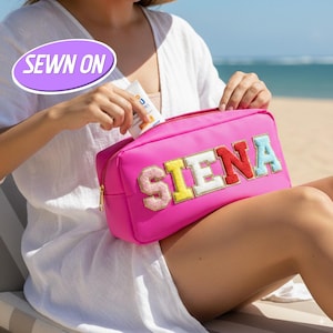Pode incluir: Uma necessaire rosa brilhante com o nome "SIENA" bordado em letras coloridas. A bolsa tem um zíper dourado e está sendo aberta para revelar um tubo de protetor solar. O fundo mostra uma cena de praia.