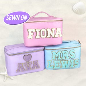 Puede incluir: Tres neceseres personalizados en rosa, lavanda y azul claro. Cada bolsa presenta un nombre o título en una fuente decorativa. La bolsa rosa dice "FIONA", la bolsa lavanda dice "AVA" y la bolsa azul dice "MRS LEWIS". Las bolsas tienen cierre de cremallera y asa superior.