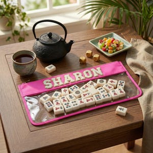 Op de afbeelding: Een houten tafel toont een zwarte theepot, een kopje thee en een roze etui met de naam "SHARON". De etui bevat witte Mahjong-tegels met rode en groene symbolen. Gouden dobbelstenen en een klein schaaltje met gekonfijte vruchten zijn ook aanwezig.