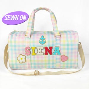 May include: A pastel plaid duffel bag with a shoulder strap and top handles. The bag features the name "SIENA" in colorful, embellished letters, along with an anchor, flower, and heart. The text "SEWN ON" is on a purple oval.