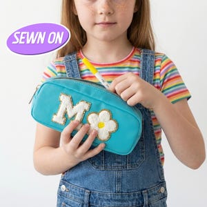 Puede incluir: Un estuche turquesa con la letra "M" blanca y dorada y una flor blanca y dorada. El estuche tiene cierre de cremallera y lo sostiene una persona que lleva una camiseta de rayas y un peto vaquero. El texto "SEWN ON" está en una burbuja morada.
