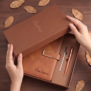 Könnte beinhalten: Braune Geschenkbox mit einem Leder-Notizbuch und Stift-Set. Das Notizbuch hat einen goldenen Verschluss, und der Name "Aaron" ist auf dem Notizbuch und der Box aufgedruckt. Eine silberne Stiftmine ist ebenfalls enthalten.
