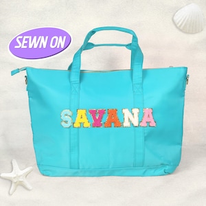 Puede incluir: Un bolso tote turquesa brillante con el nombre "SAVANA" en letras coloridas cosidas. El bolso tiene cierre de cremallera y asas. Un óvalo morado con las palabras "SEWN ON" está en la esquina superior izquierda. Una estrella de mar y una concha están en el fondo.