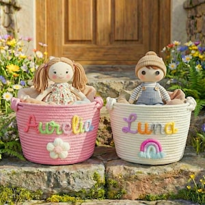 May include: Two decorative rope baskets, one pink and one cream, each containing a doll. The pink basket has the name "Aurelia" and a flower, while the cream basket has "Luna" and a rainbow. The dolls are dressed in fabric clothing.