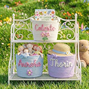 May include: A white metal shelf unit displays three rope baskets. The top basket is white with the name "Celeste" and a book titled "My Garden Friends." The middle basket is blue with the name "Aurora." The bottom basket is purple with the name "Thorin."