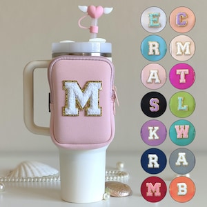 Könnte beinhalten: Ein rosa isolierter Becher mit Griff und einer passenden rosa Tasche mit dem goldverzierten weißen Buchstaben "M". Der Becher hat einen herzförmigen Strohhalmaufsatz mit Flügeln. Zusätzliche Buchstabenaufnäher in verschiedenen Farben werden angezeigt.