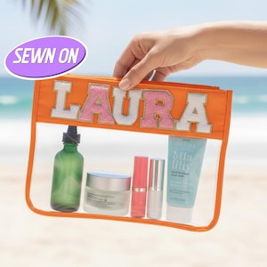 Puede incluir: Un neceser transparente con una sección superior naranja que muestra el nombre "LAURA" en letras blancas y rosas. La bolsa contiene productos de belleza como una botella verde, un tarro, barras de labios y un tubo de loción. Las palabras "SEWN ON" están en un óvalo morado.
