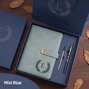 May include: A mist blue journal and pen set presented in a gift box. The journal features a gold clasp and a laurel wreath with the letter "E". A matching pen and a silver pen are included. The gift box and bag are also mist blue.