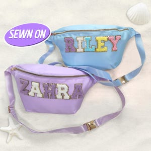 May include: Two personalized fanny packs. One is light blue with the name "RILEY" in colorful sequined letters. The other is lavender with "ZAHRA" in sequined letters. Both have gold zippers and adjustable straps.