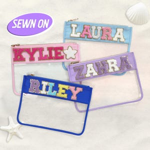 May include: Four personalised clear pencil cases with names in colourful, glittery letters. The cases are in blue, pink, and lavender, with gold zips. The text "SEWN ON" is in a purple oval.
