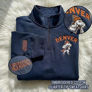 May include: Navy blue embroidered cotton quarter zip sweatshirt. Features the word "DENVER" in orange above a cartoon horse holding a football. The sleeve has the number "5280" embroidered in orange. The sweatshirt has a zipper and a high collar.