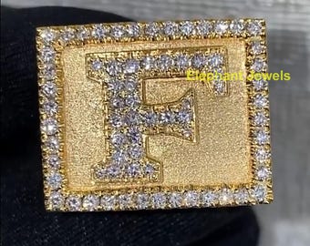May include: A gold-colored ring with the letter "F" encrusted with small, sparkling diamonds. The ring has a rectangular shape with a border of additional diamonds. The text "Elephant Jewels" is visible.