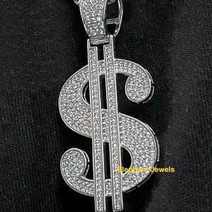 May include: A silver-colored pendant in the shape of a dollar sign, encrusted with small, sparkling stones. The pendant hangs from a silver chain. The text "Elephant Jewels" is visible on the pendant.
