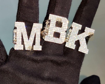 May include: Three diamond-encrusted rings spelling out "MBK" are displayed. The rings are set against a black background. Each letter is fully covered in small, sparkling diamonds, creating a luxurious and eye-catching design. The rings are a statement piece.