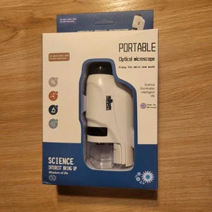 Peut inclure: Un microscope optique portable blanc et noir dans son emballage d'origine. La boîte est blanche et bleue avec les mots "PORTABLE Optical microscope" et "SCIENCE INTEREST BRING UP" imprimés dessus. Le microscope est visible à travers une fenêtre transparente.