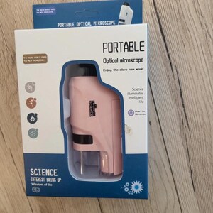 Peut inclure: Un microscope optique portable rose dans son emballage. La boîte est blanche et bleue, avec les mots "PORTABLE Optical microscope" imprimés sur le devant. Le texte "SCIENCE INTEREST BRING UP Wisdom of life" est également visible.
