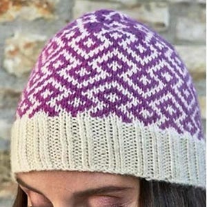May include: A knit beanie featuring a purple and white geometric pattern across the crown and a solid cream-colored ribbed brim. The hat is constructed with a dense, textured knit stitch, showcasing a repeating diamond-like motif throughout.