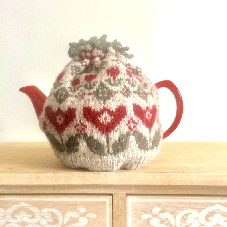 May include: A red teapot with a knitted cozy featuring a floral pattern in shades of beige, gray, and red. The cozy has a pom-pom on top.
